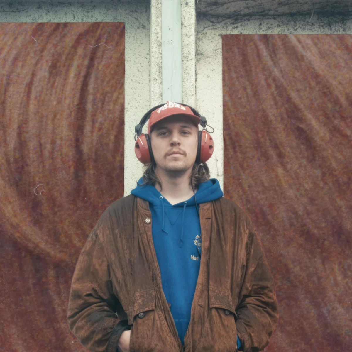 Person in a red cap and large red headphones, wearing a blue hoodie and brown jacket against a textured red backdrop.