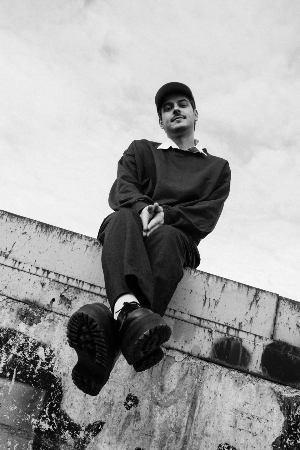 Black and white low-angle shot of a person in a cap and sweatshirt sitting on a concrete ledge against a cloudy sky.