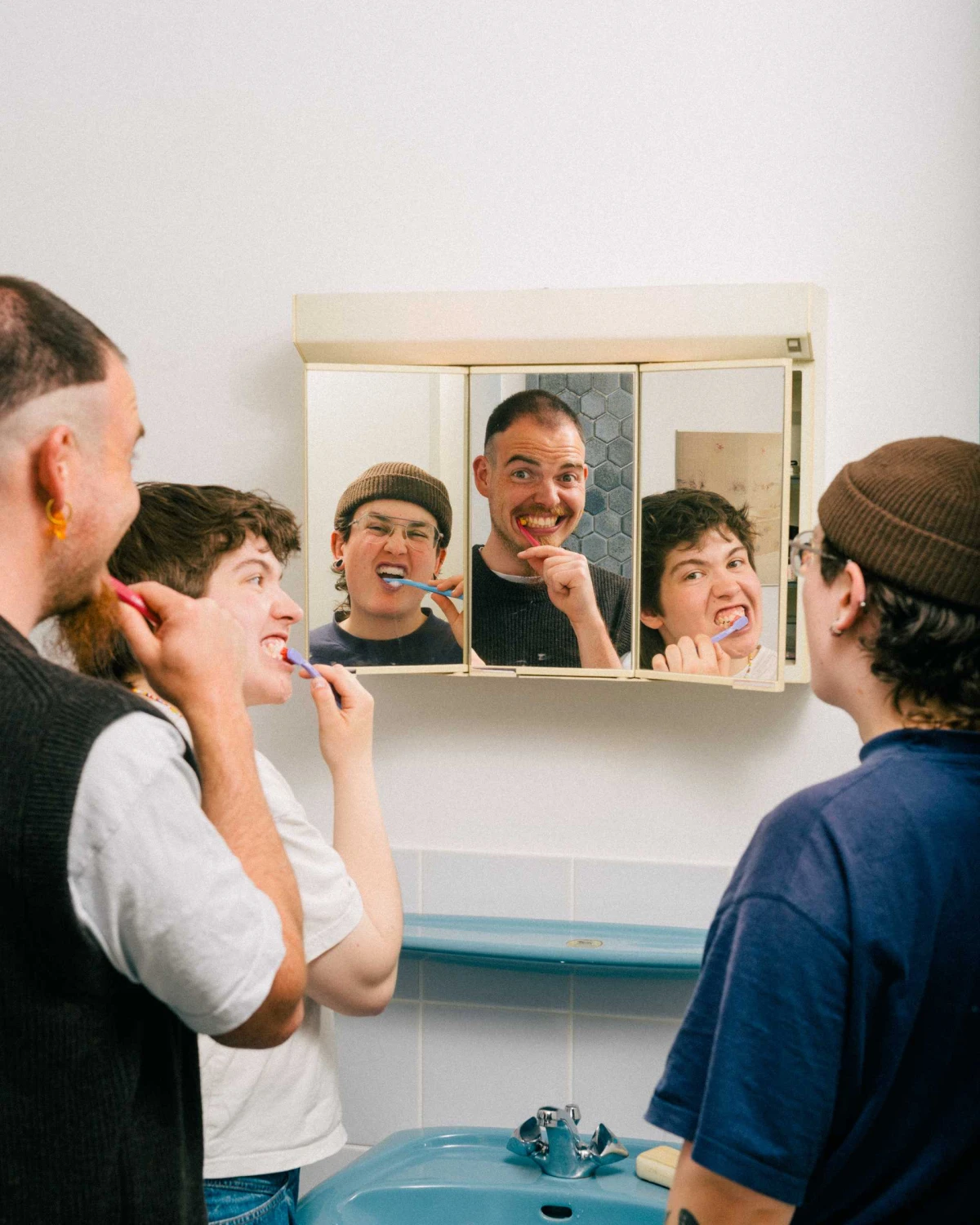 Three people brush their teeth together, their reflections visible in a three-paneled bathroom mirror cabinet.