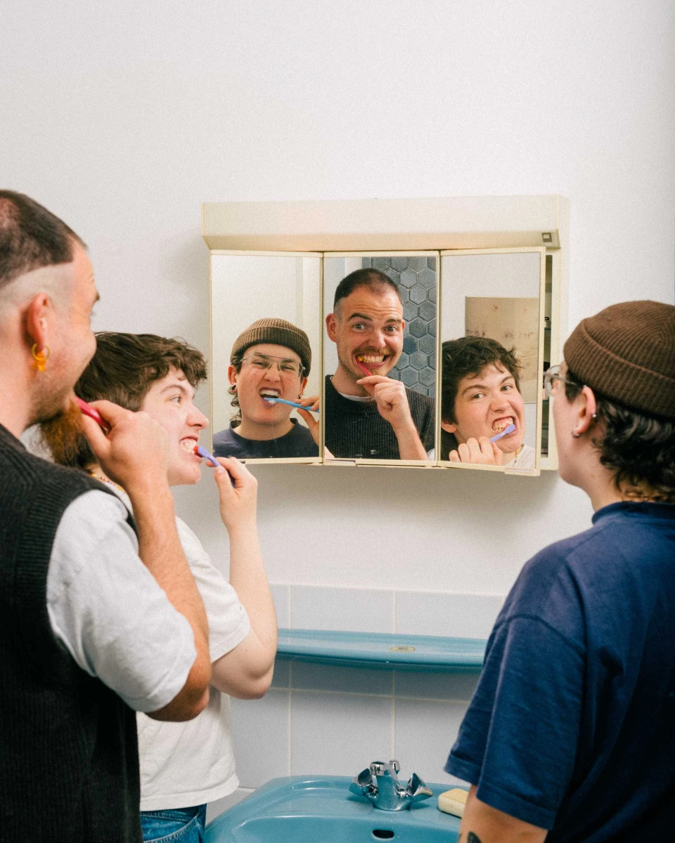 Three people brush their teeth together, their reflections visible in a three-paneled bathroom mirror cabinet.