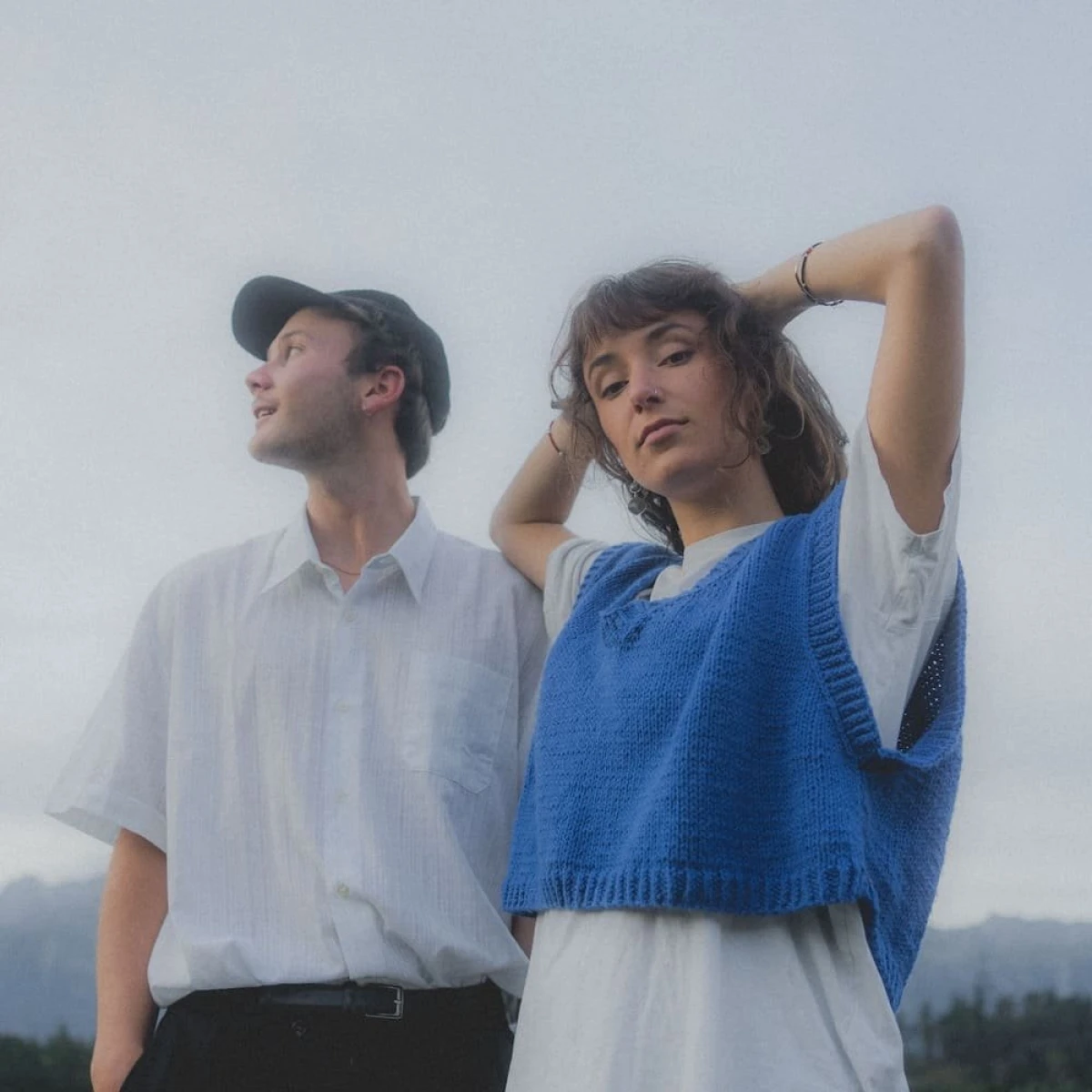 Two people standing outdoors against a pale sky; one in a blue knit vest and one in a white shirt and black cap.