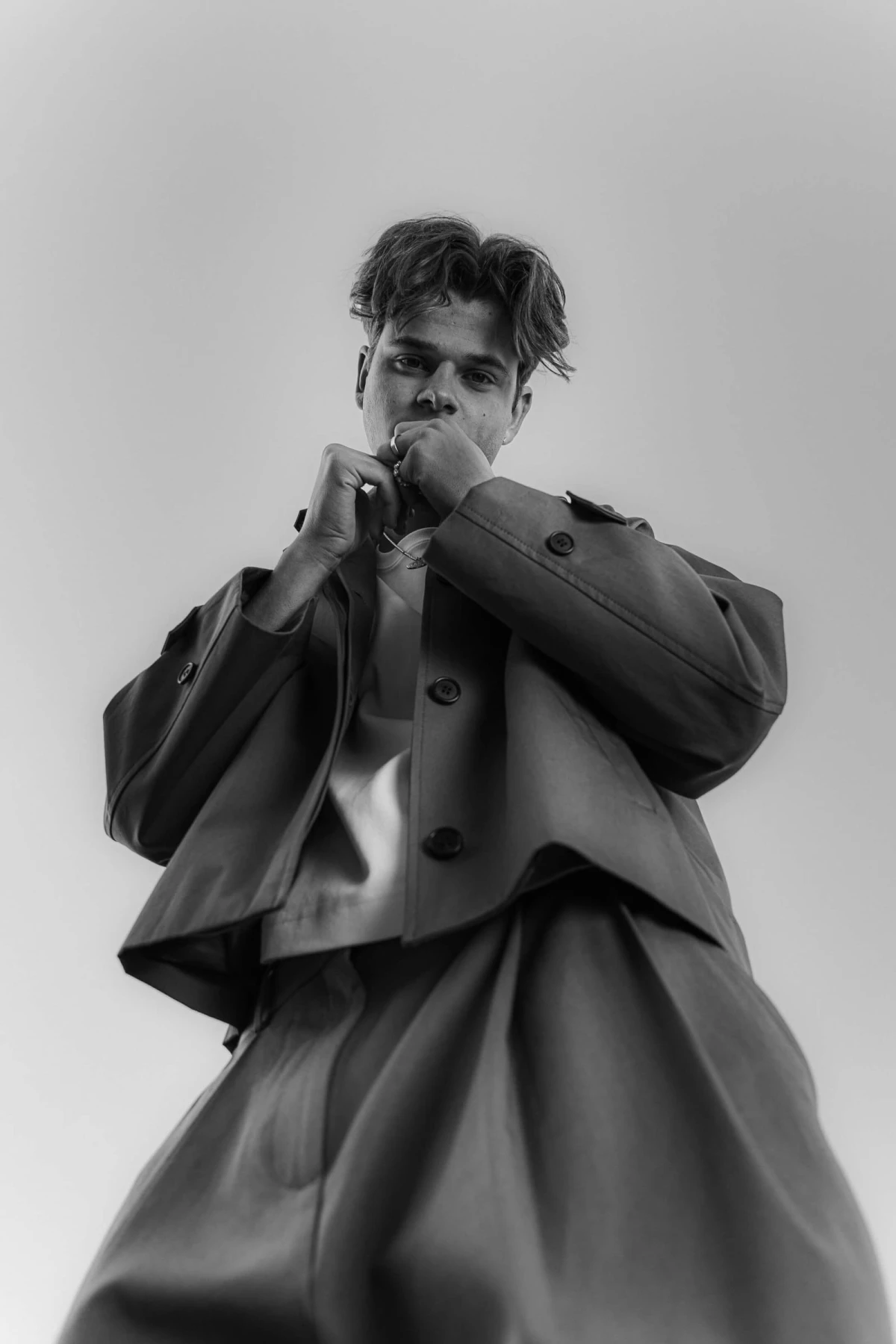 Black and white low-angle shot of a person in a wide-cut jacket and trousers, holding their hands to their face.