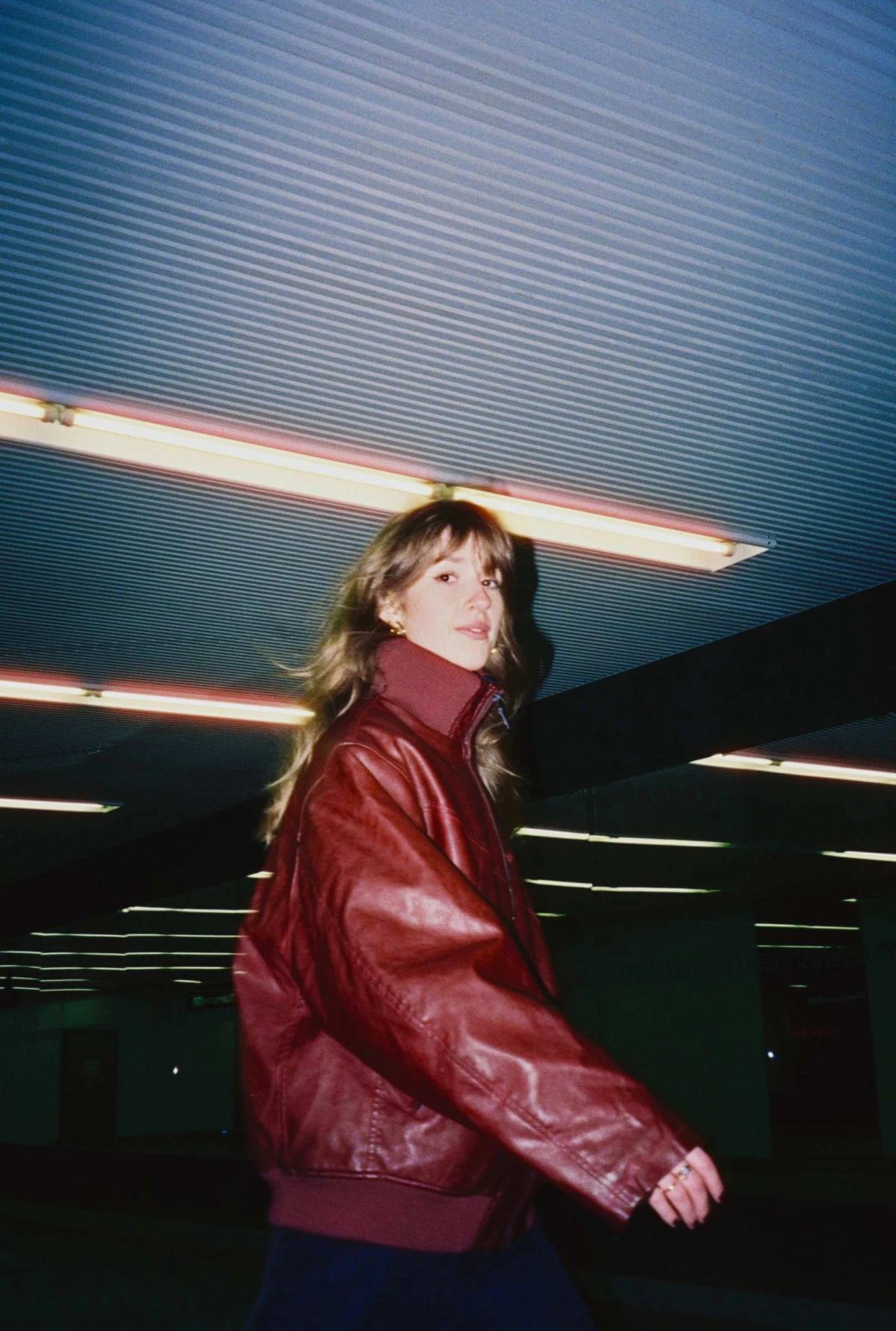 Person in a maroon leather bomber jacket walking under bright fluorescent tube lights in a dark, ribbed ceiling space.