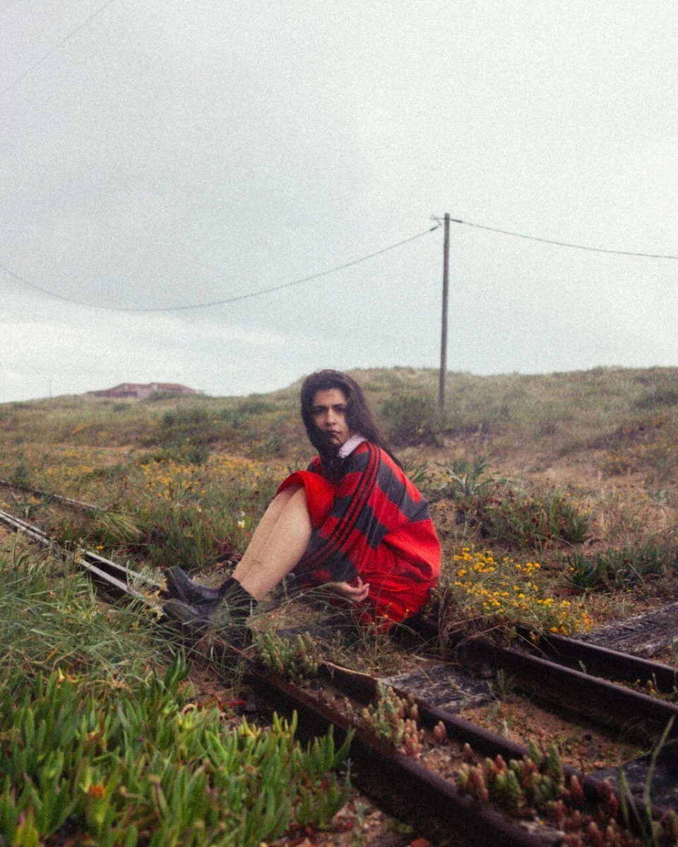 Person in a red and black striped shirt sitting on abandoned train tracks in a grassy, overcast field.