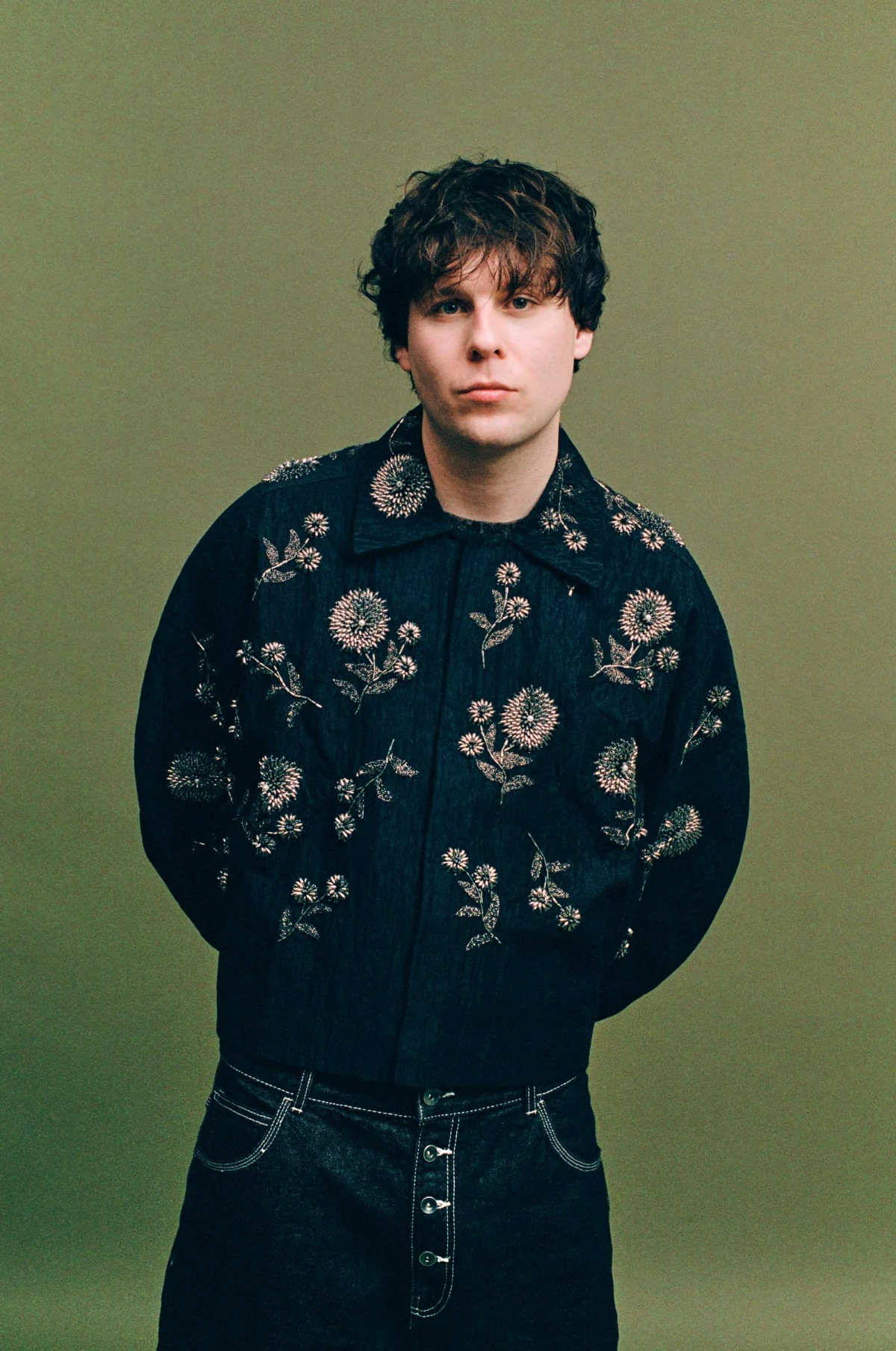 Person with dark curly hair wearing a black jacket with floral embroidery, posing against an olive green background.