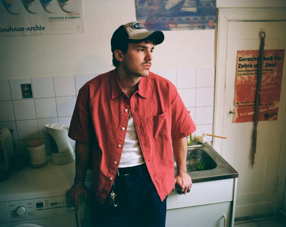 Person with a mustache and cap wearing a red plaid shirt, leaning against a kitchen sink and washing machine.