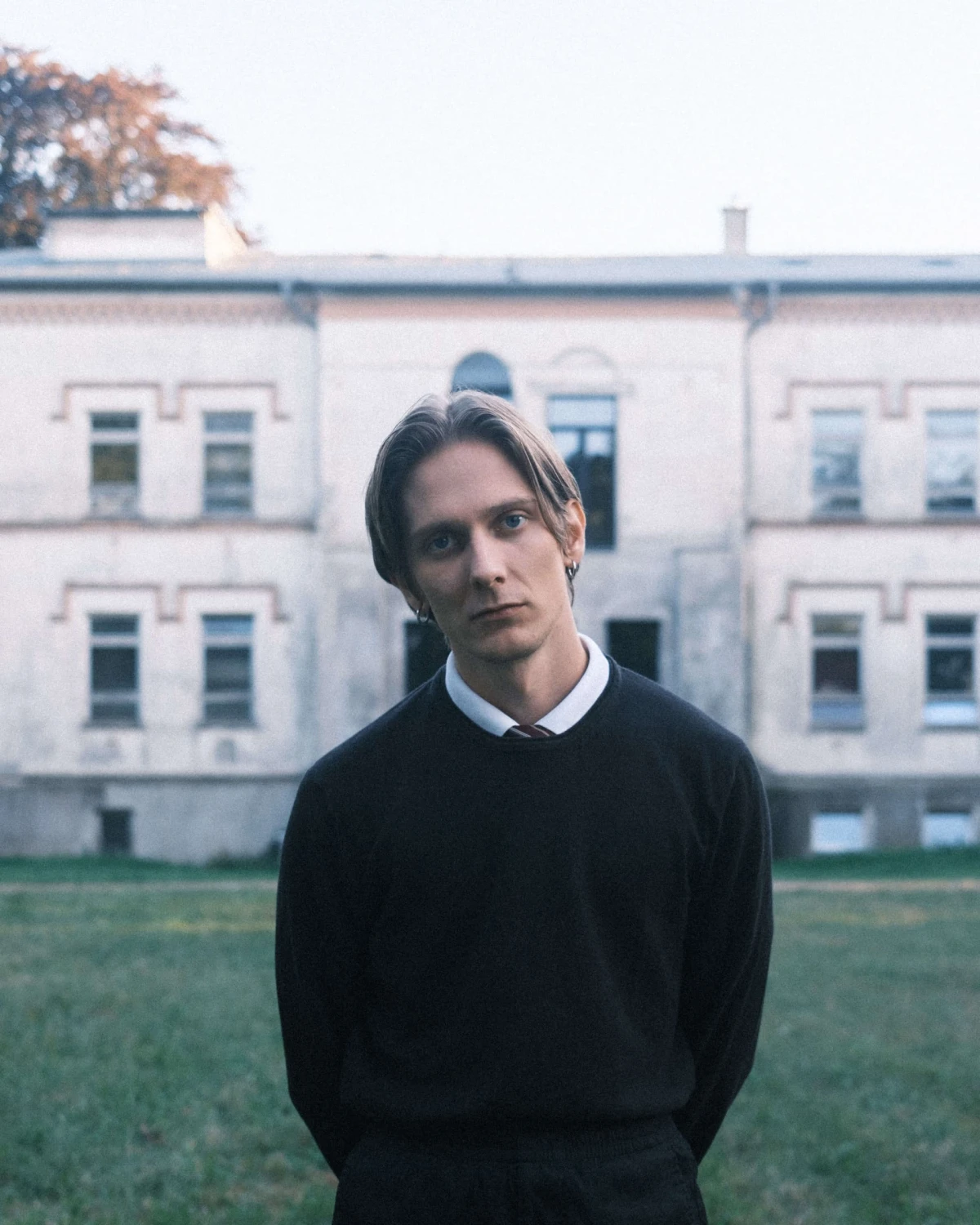 Person in a black sweater and collared shirt standing in front of a large, pale building with many windows.