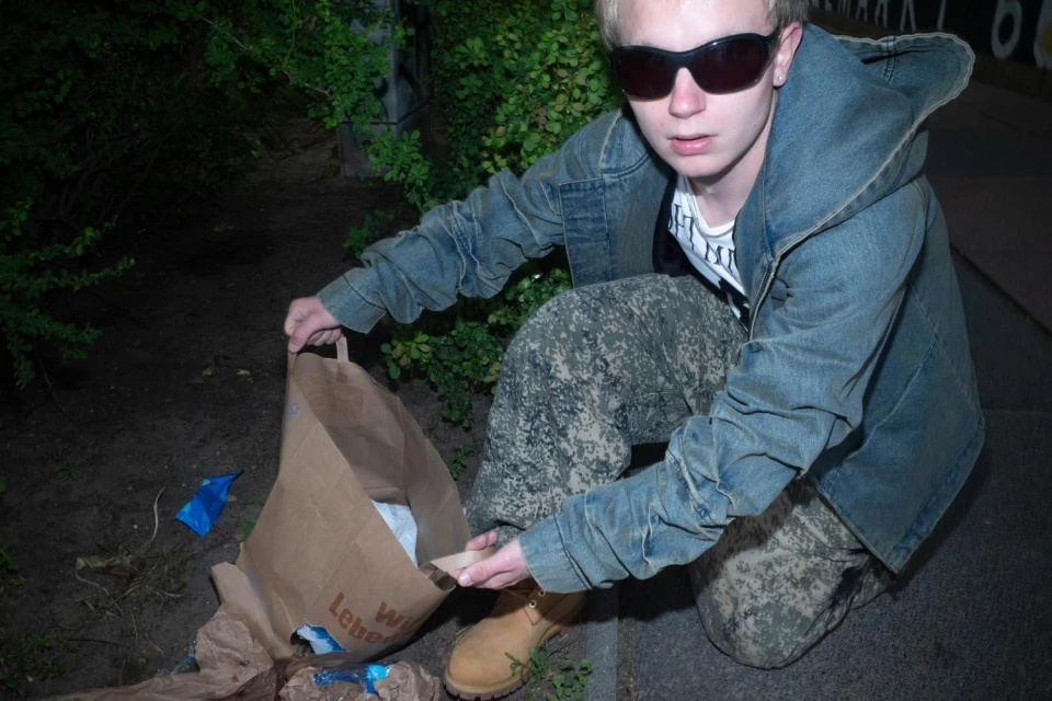 Person in sunglasses, a denim jacket, and camouflage pants kneels outdoors holding a brown paper bag at night.
