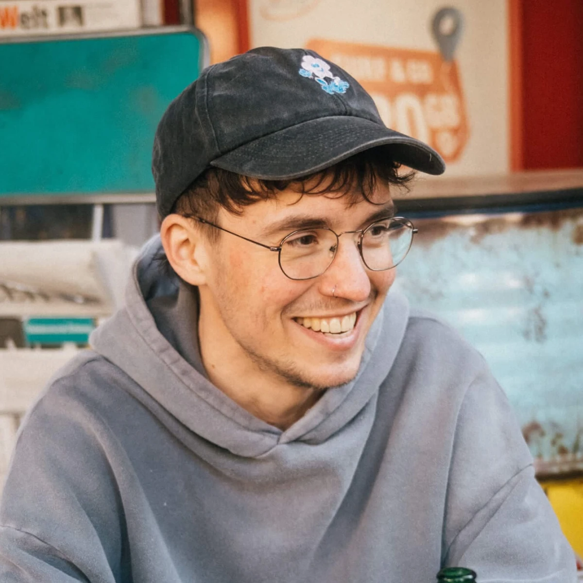 Smiling person in a grey hoodie, glasses, and cap sits at a table with a bottle near a newspaper rack.