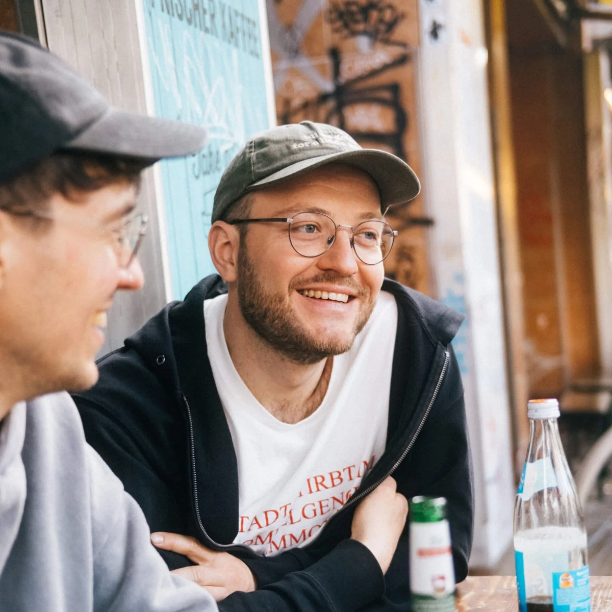 Smiling person with a beard, glasses, and green cap sitting at a wooden table in an urban outdoor setting.