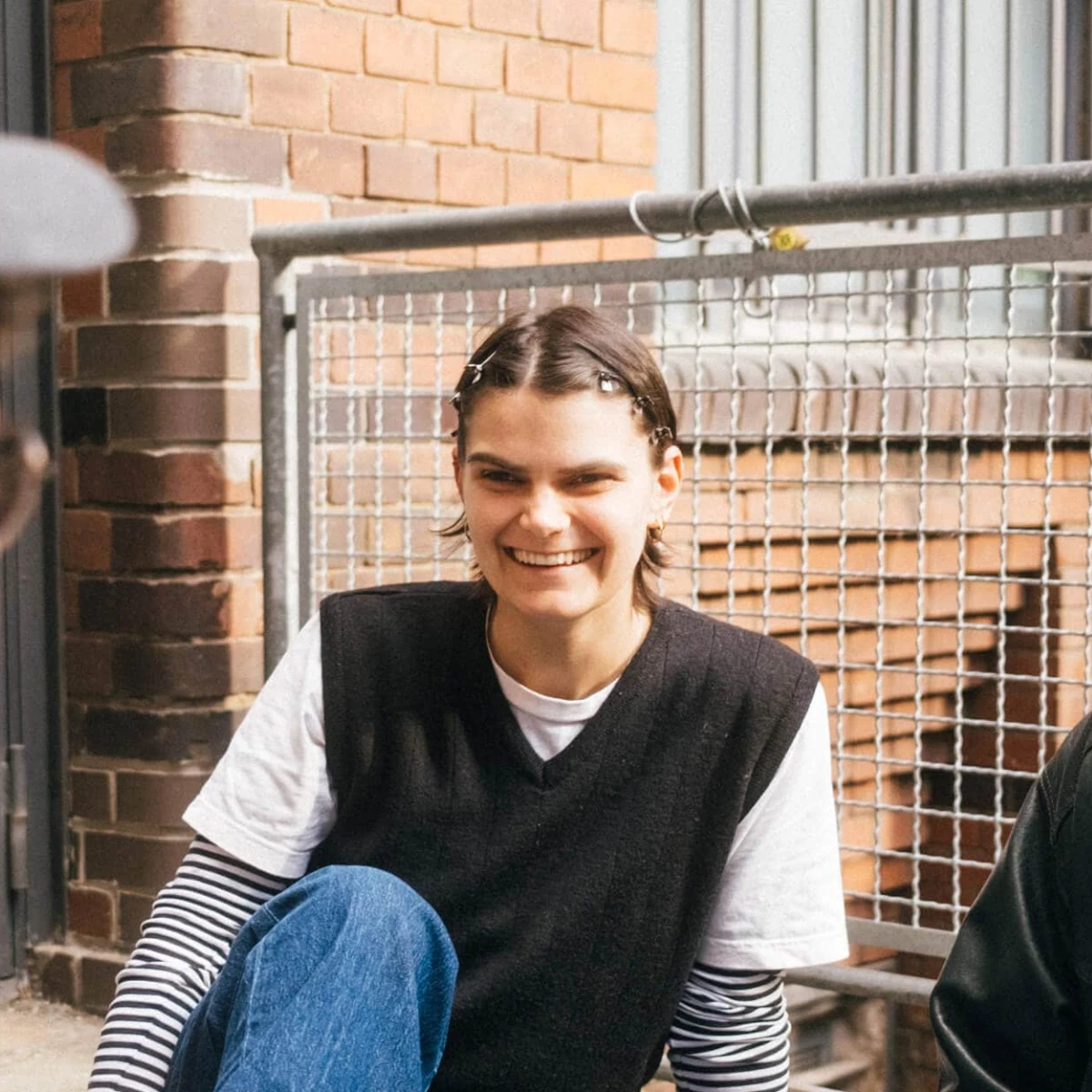 Smiling person in a black vest and striped shirt sits outdoors with others on the ground near a brick wall.